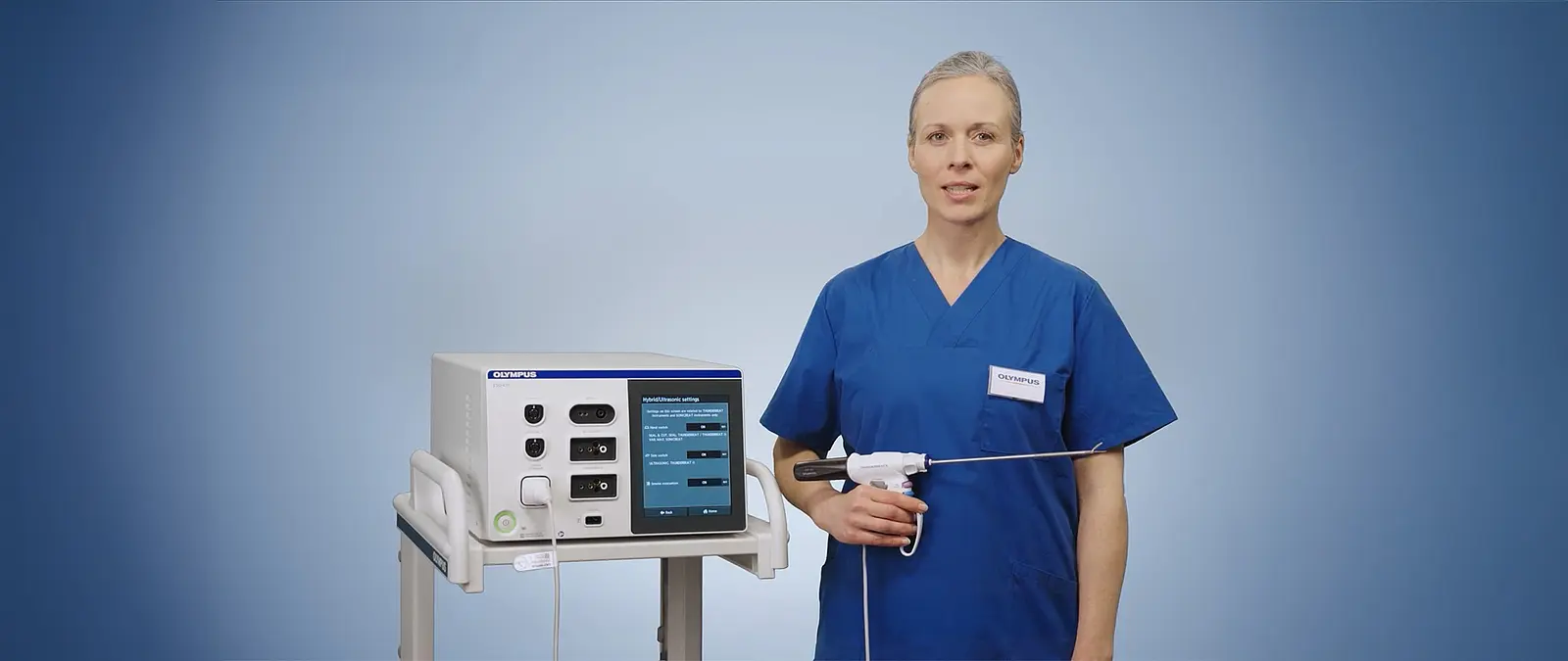 Eine medizinische Fachkraft in blauer OP-Kleidung demonstriert das Olympus THUNDERBEAT II System neben einem ESG-410 Generator vor neutralem Hintergrund.