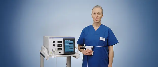 Eine medizinische Fachkraft in blauer OP-Kleidung demonstriert das Olympus THUNDERBEAT II System neben einem ESG-410 Generator vor neutralem Hintergrund.