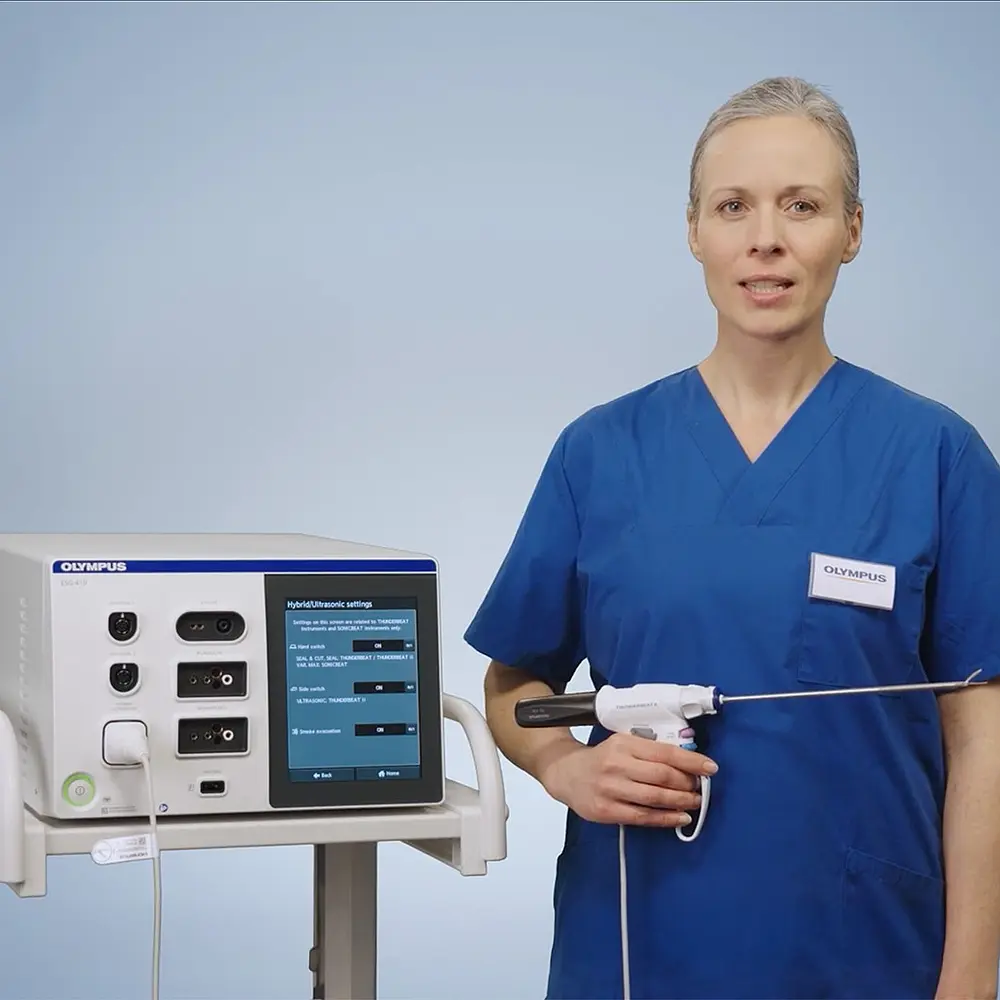 Eine medizinische Fachkraft in blauer OP-Kleidung demonstriert das Olympus THUNDERBEAT II System neben einem ESG-410 Generator vor neutralem Hintergrund.