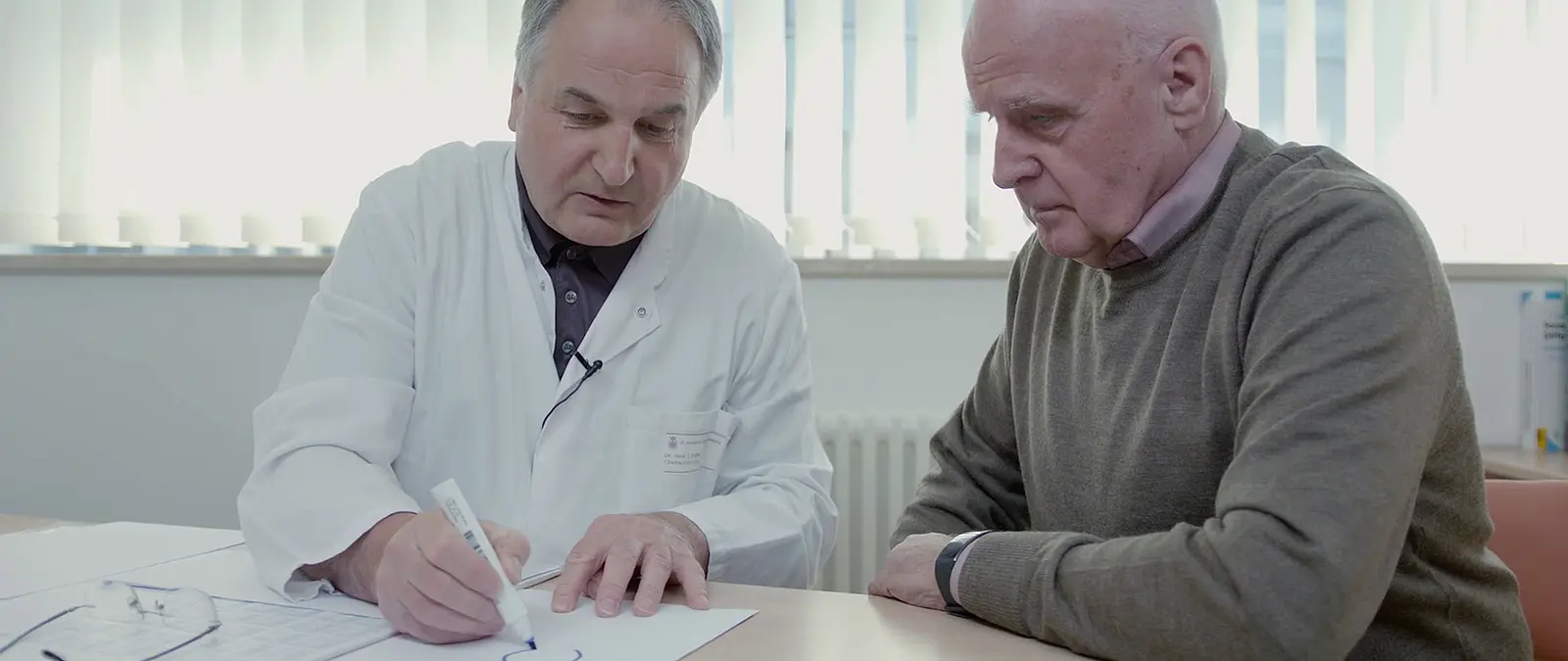 Arzt erklärt einem älteren Patienten etwas am Tisch im Olympus Dokumentarfilm