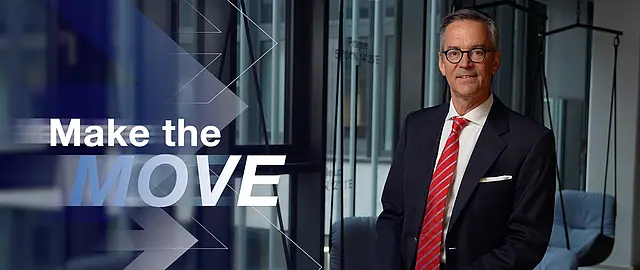 Geschäftsmann im Anzug steht in einem modernen Innenraum vor großem Schriftzug „Make the MOVE“ – Keyvisual für Testimonial-Video und vertrauensvolle Markenkommunikation im medizinischen Bereich.