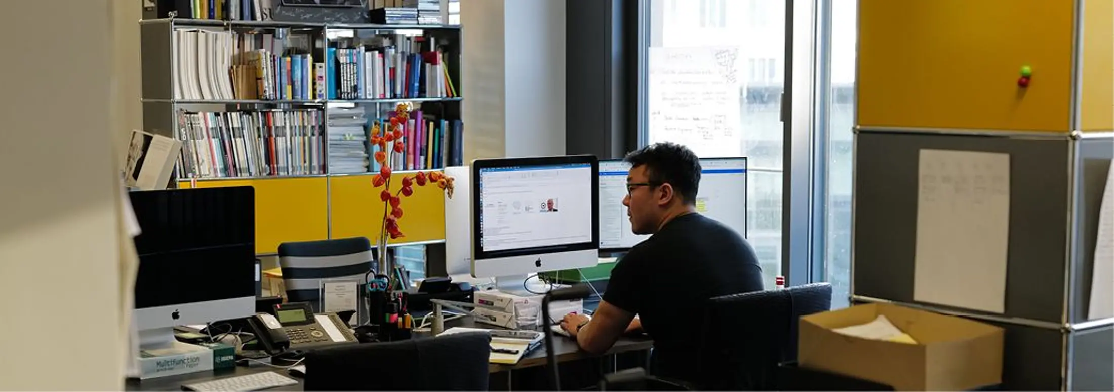 Ein Mitarbeiter sitzt in einem Büro an einem Schreibtisch und arbeitet an zwei Computerbildschirmen. Im Hintergrund stehen Bücherregale, Büromaterialien und gelbe Regalelemente; die Szene wirkt wie ein Arbeitsplatz in der Softwareentwicklung.