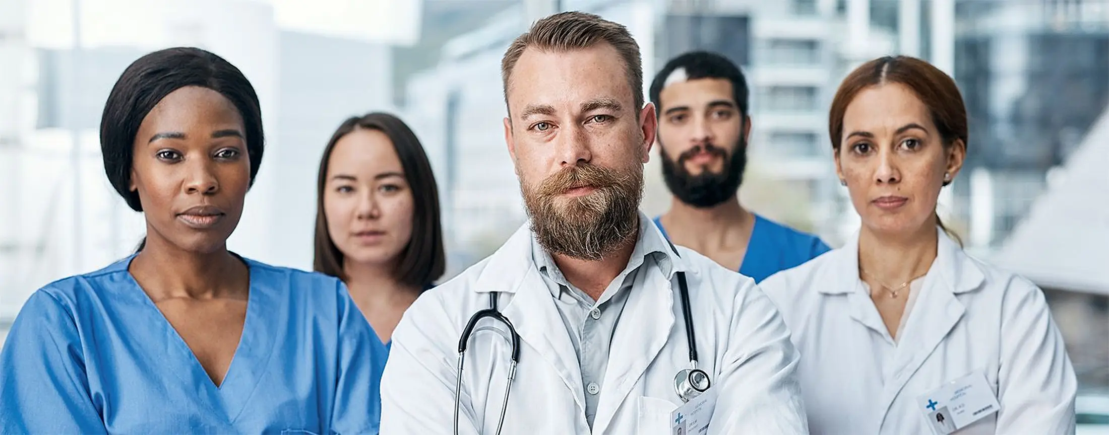 Ein interprofessionelles Team aus Ärztinnen, Ärzten und Pflegekräften steht in einem hellen Krankenhausflur und blickt selbstbewusst in die Kamera. Im Vordergrund tragen zwei Personen weiße Kittel, weitere Mitarbeitende tragen blaue medizinische Kleidung.
