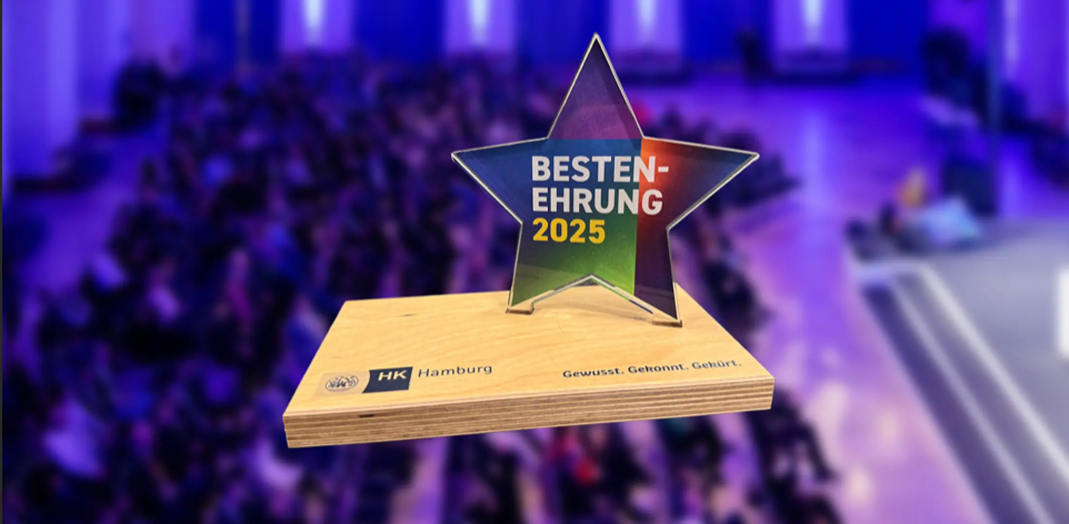 Ein sternförmiger Preis der Handelskammer Hamburg mit der Aufschrift „Bestenehrung 2025“ steht auf einem Holzsockel. Im unscharfen Hintergrund ist eine festlich beleuchtete Veranstaltung mit Publikum in blau-violettem Licht zu sehen.