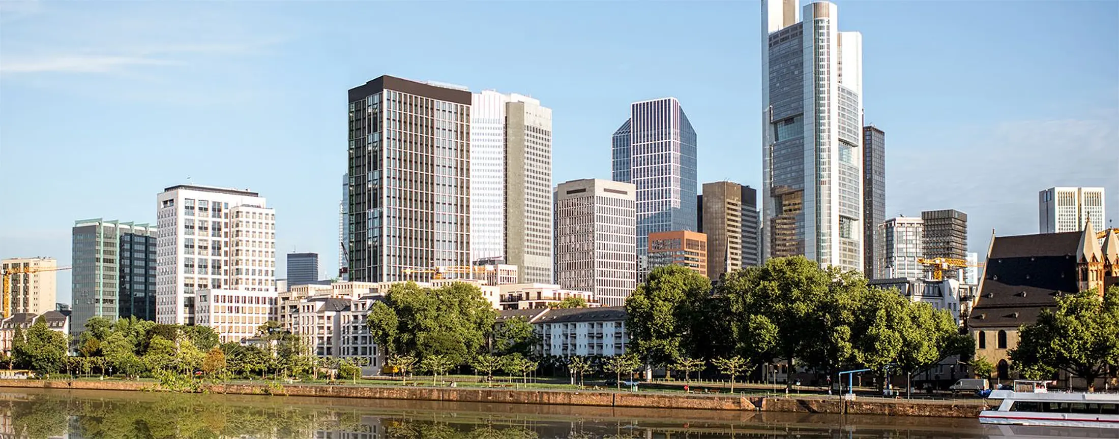 Büro- und Bank-Gebäude stehen hinter einer Reihe grüner Bäume am Mainufer in Frankfurt