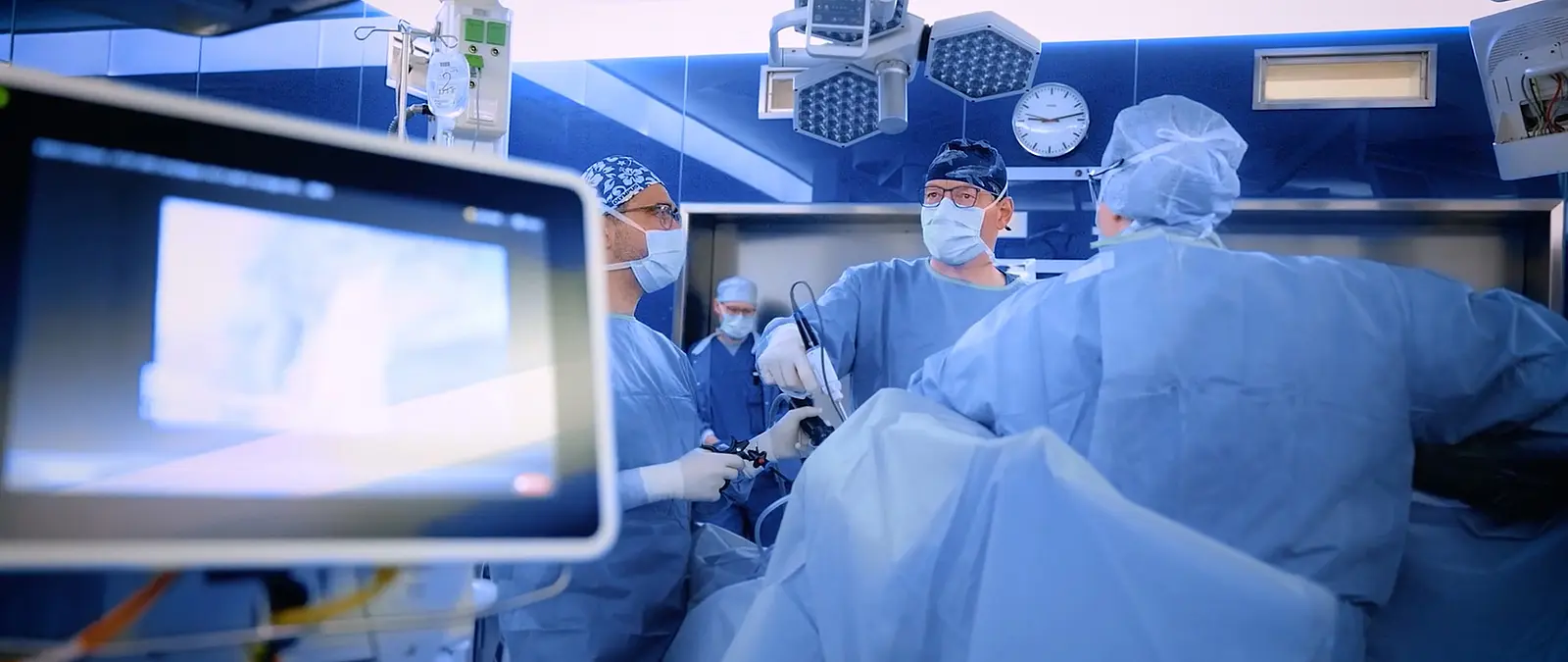 Ein OP-Team in voller Schutzkleidung führt eine minimalinvasive Operation durch. Im Vordergrund ist ein Bildschirm mit Endoskopiebild zu sehen. Verwendet werden chirurgische Geräte aus dem Olympus Surgery Portfolio wie THUNDERBEAT oder ENDOEYE.