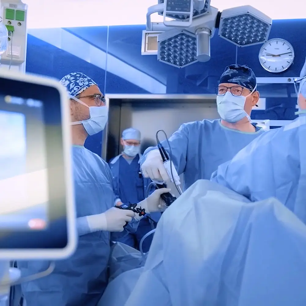 Ein OP-Team in voller Schutzkleidung führt eine minimalinvasive Operation durch. Im Vordergrund ist ein Bildschirm mit Endoskopiebild zu sehen. Verwendet werden chirurgische Geräte aus dem Olympus Surgery Portfolio wie THUNDERBEAT oder ENDOEYE.