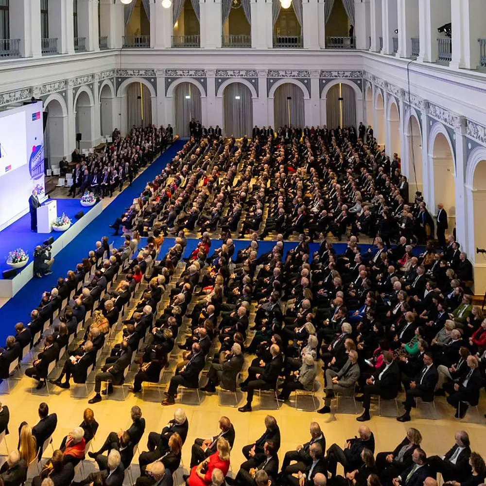 Große Konferenz mit hunderten Teilnehmenden in einem historischen Saal, Blick von oben auf ein voll besetztes Auditorium mit Bühne, Rednerpult und Leinwand