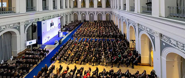 Große Konferenz mit hunderten Teilnehmenden in einem historischen Saal, Blick von oben auf ein voll besetztes Auditorium mit Bühne, Rednerpult und Leinwand