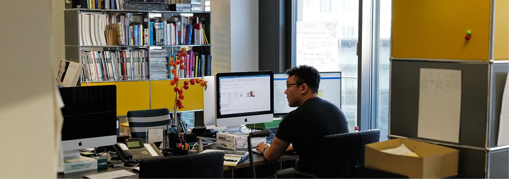 Mitarbeiter in einem Büro am Schreibtisch vor zwei Monitoren, mit Bücherregal und gelben Schrankelementen im Hintergrund.